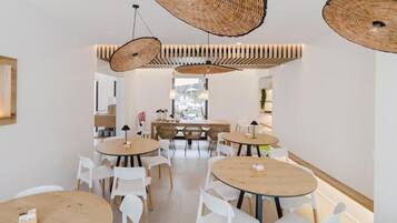 A modern dining area with round tables and white chairs, featuring woven ceiling light fixtures and a view through a glass door.