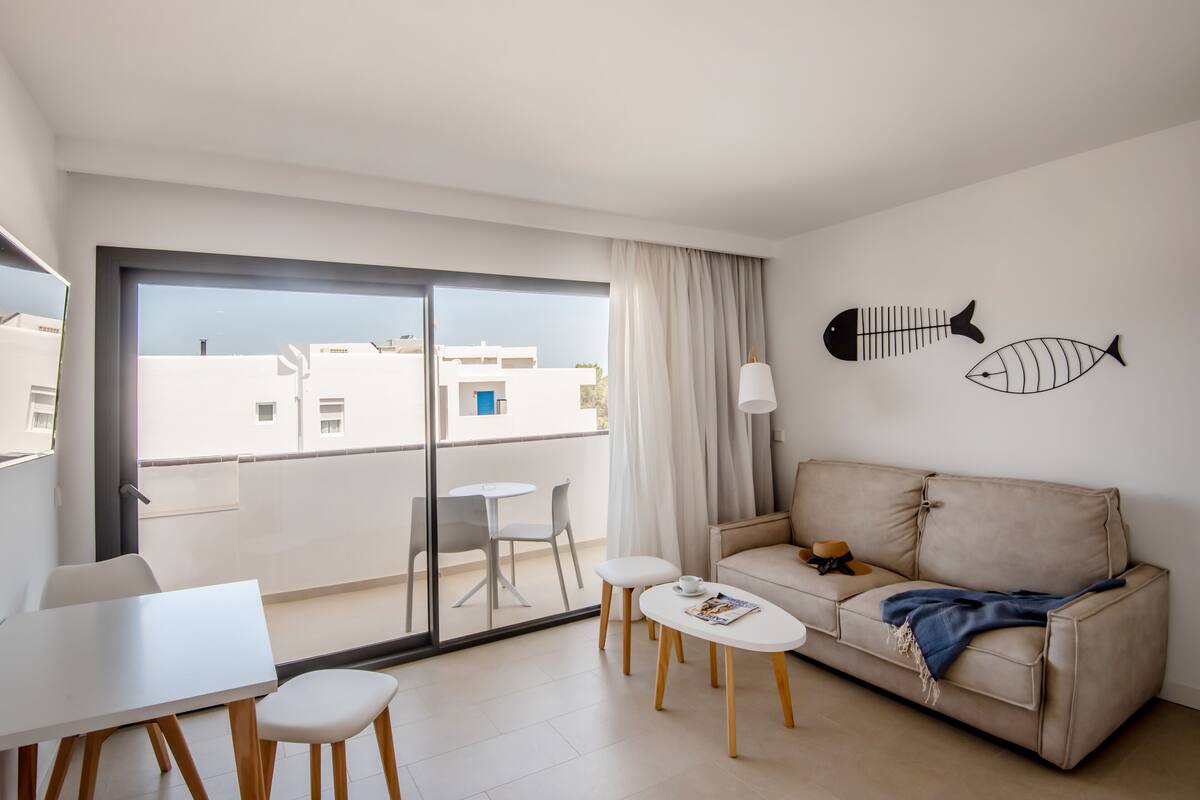 A modern living room with a sofa, coffee table, and chairs, a balcony with a view of buildings, and two wall-mounted fish decorations.