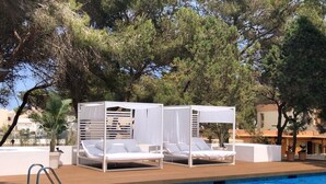 Uma área à beira da piscina com duas espreguiçadeiras brancas sob um toldo, uma piscina azul cristalina e árvores ao fundo.