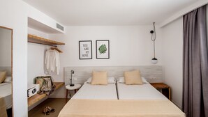 Quarto de hotel com uma cama, mesa de cabeceira, espelho, prateleira com roupas e malas, e duas imagens emolduradas na parede.