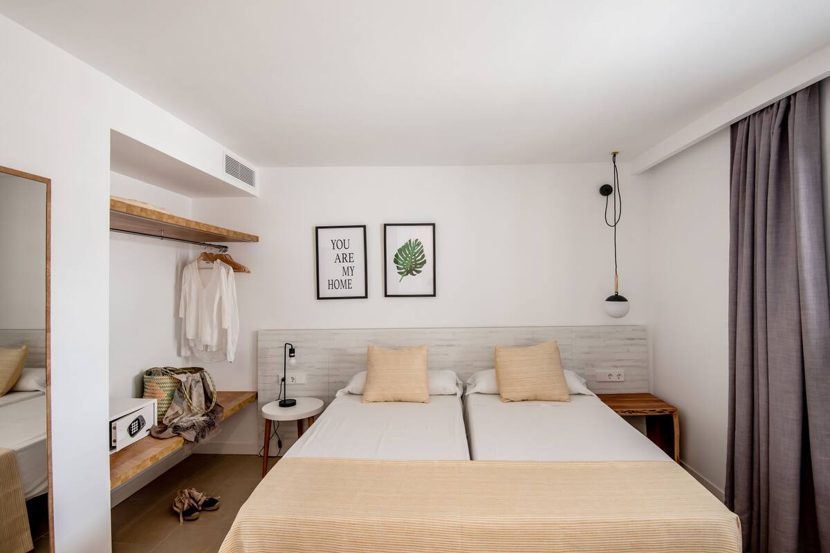 A hotel room with a bed, a bedside table, a mirror, a shelf with clothes and bags, and two framed pictures on the wall.