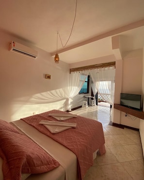 Un dormitorio con cama, televisión y ventana con cortinas traslúcidas.
