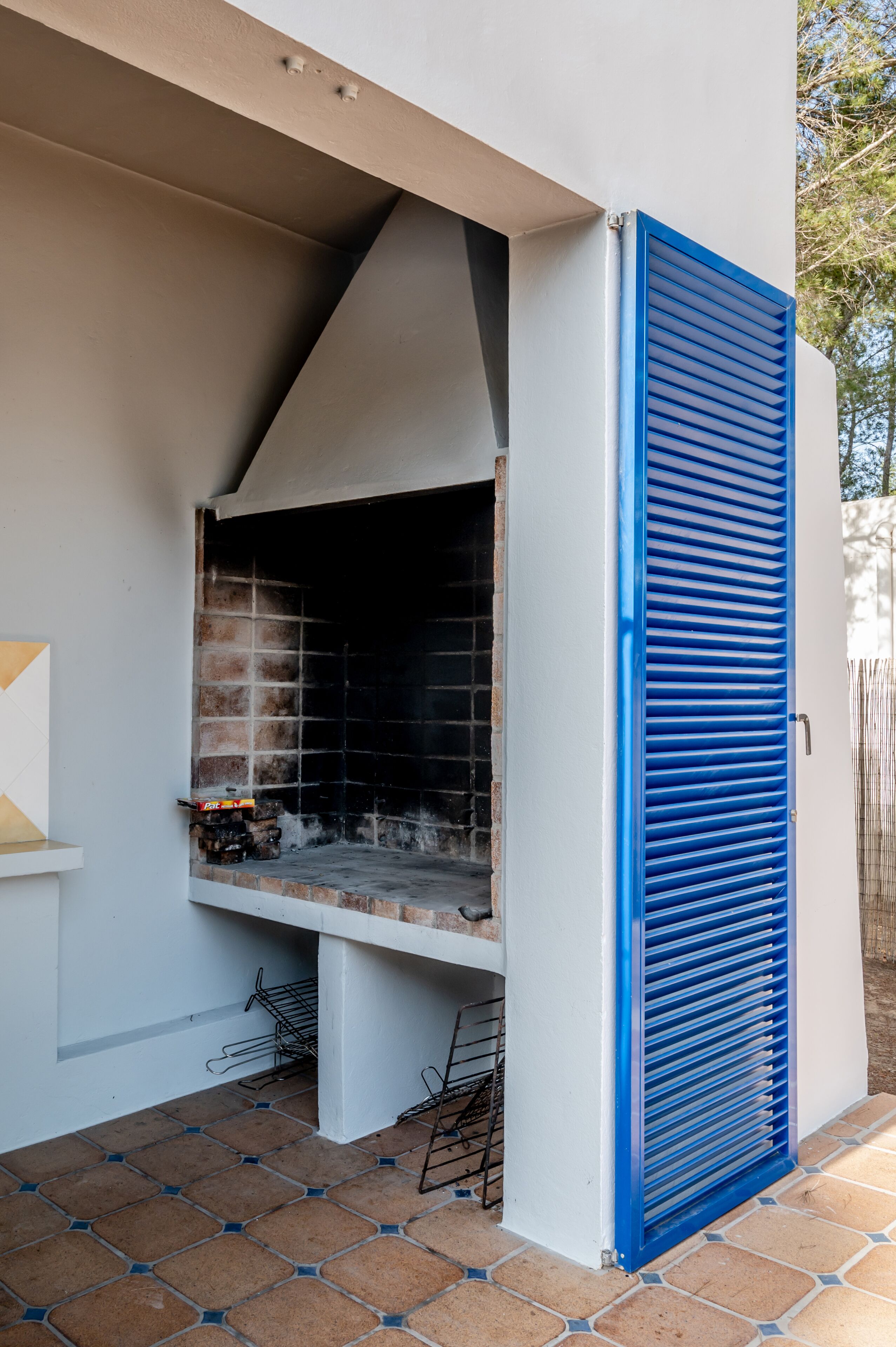 An outdoor grill area with a white wall and blue shutters, a tiled patio, and a brick fireplace.
