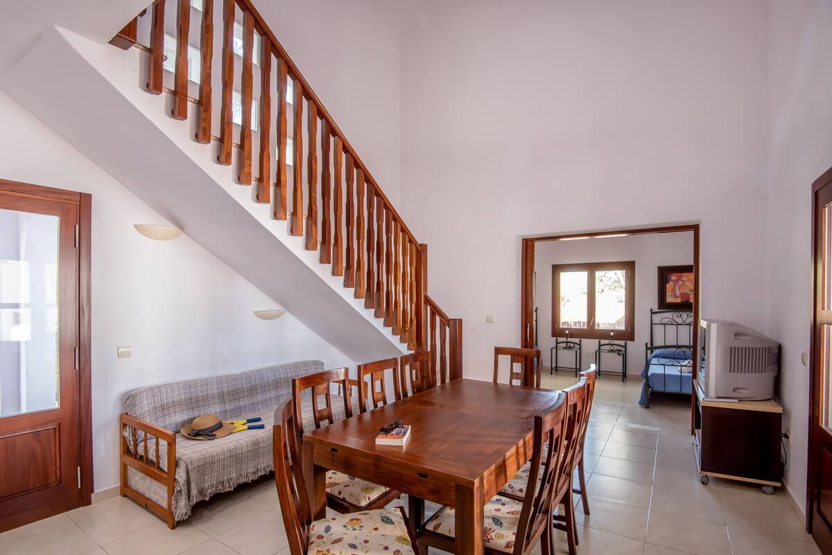 A spacious dining area with a wooden table and chairs, a staircase, a sofa, and a television in the corner.