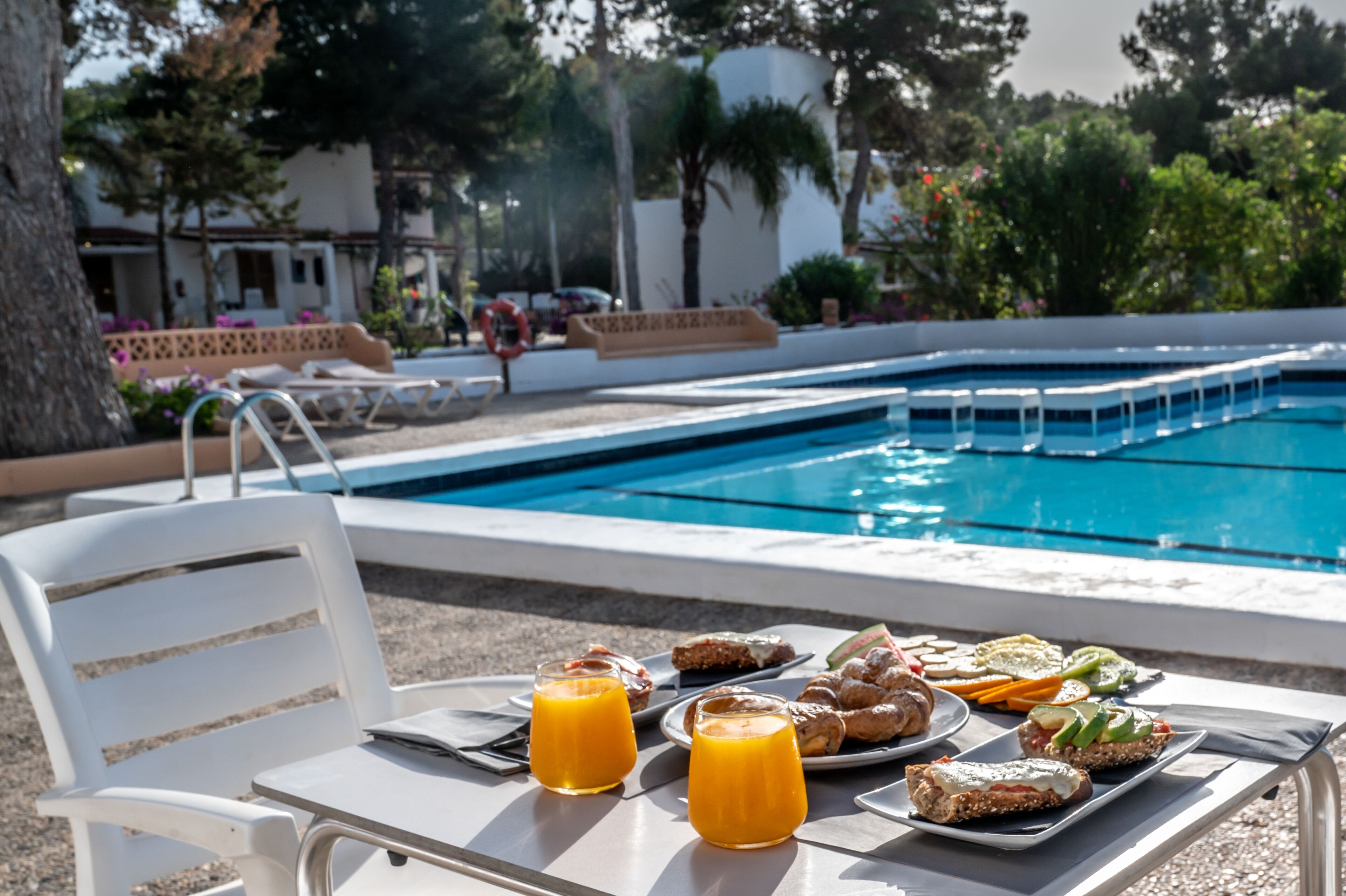 A poolside breakfast setup with a table, chairs, and a variety of food and drinks.