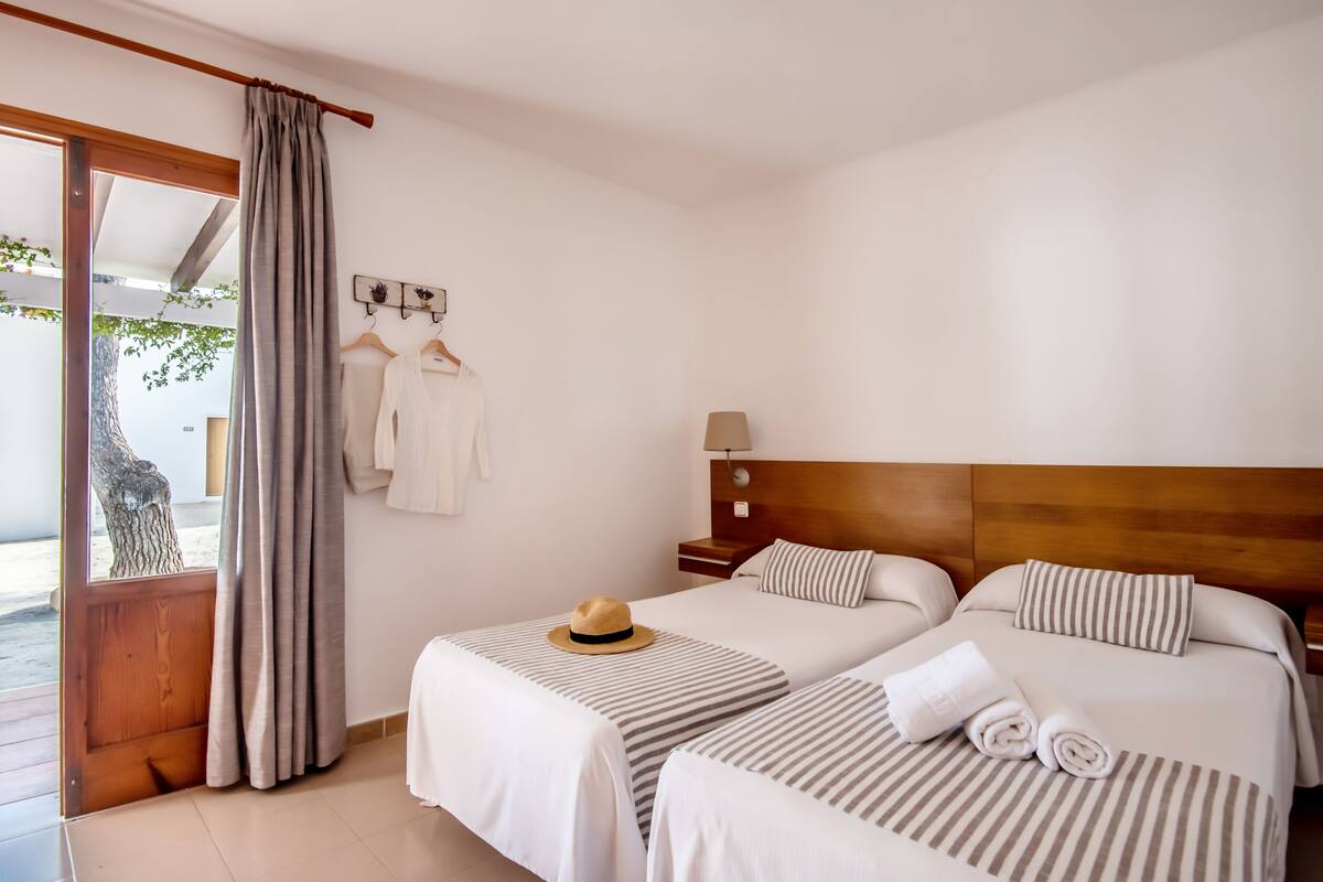 A hotel room with two beds, a wooden headboard, a nightstand, a lamp, and a view of the outdoors.