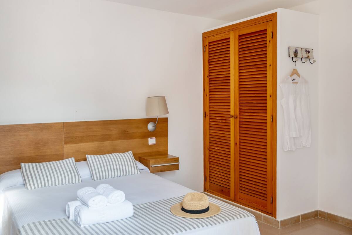 A neatly made bed with striped bedding, a bedside lamp, and a hat on the bed.