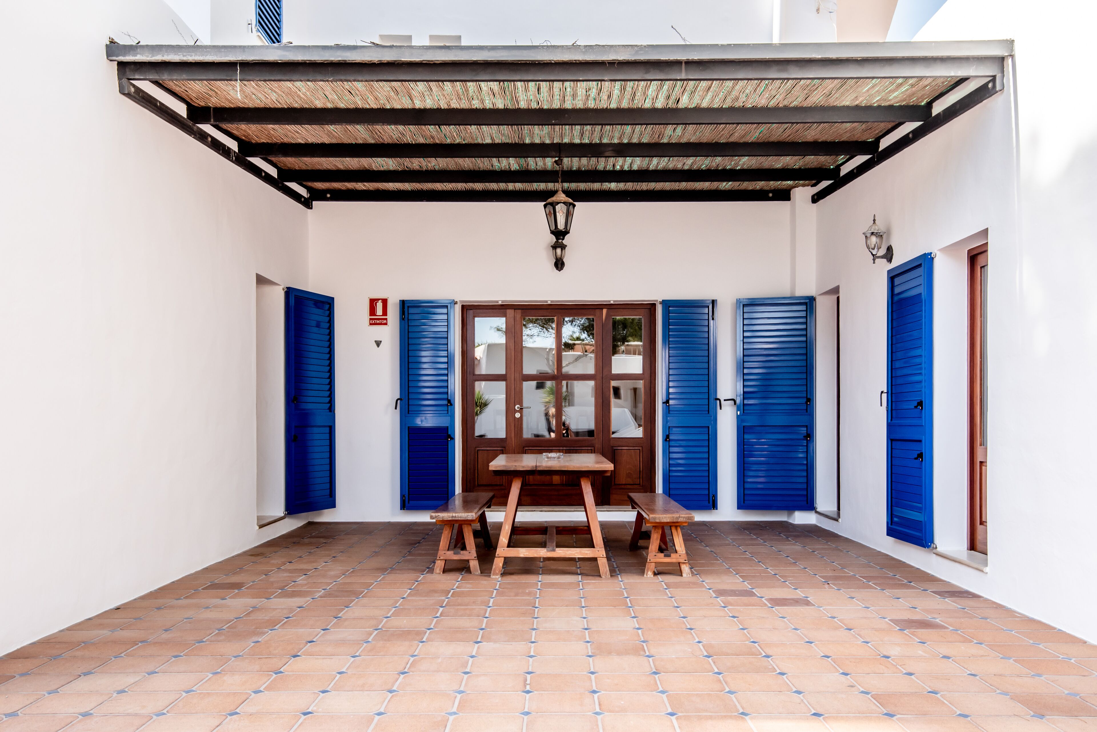 A covered patio with a wooden table and benches, a door with blue shutters, and a tiled floor.