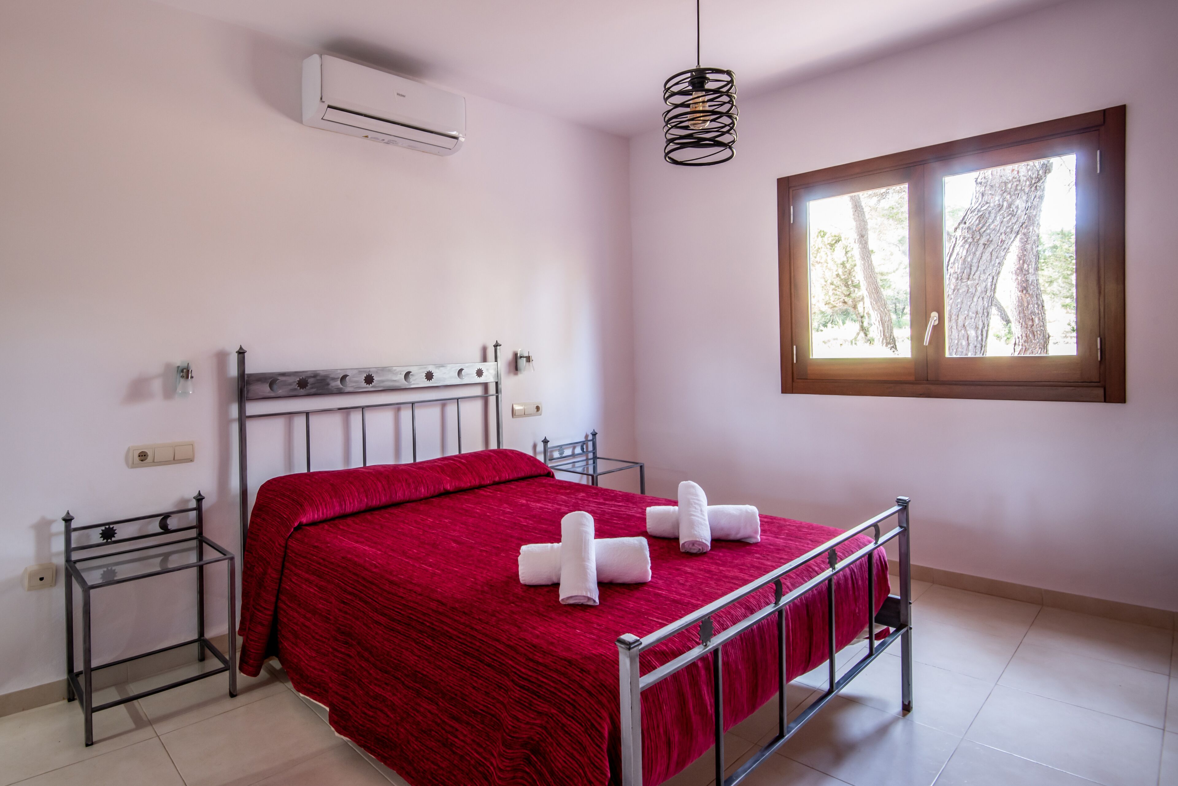 Ein Einzelbett mit roter Bettdecke und weißen Handtüchern, ein Metall-Kopfteil, ein kleiner Nachttisch, eine Wand-Klimaanlage, eine Pendelleuchte und ein Fenster mit Blick auf Bäume.