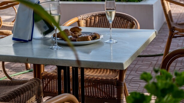 A table with a glass of water and a plate of pastries, wicker chairs, and a pool in the background. - Hostal Es Pi (Formentera)