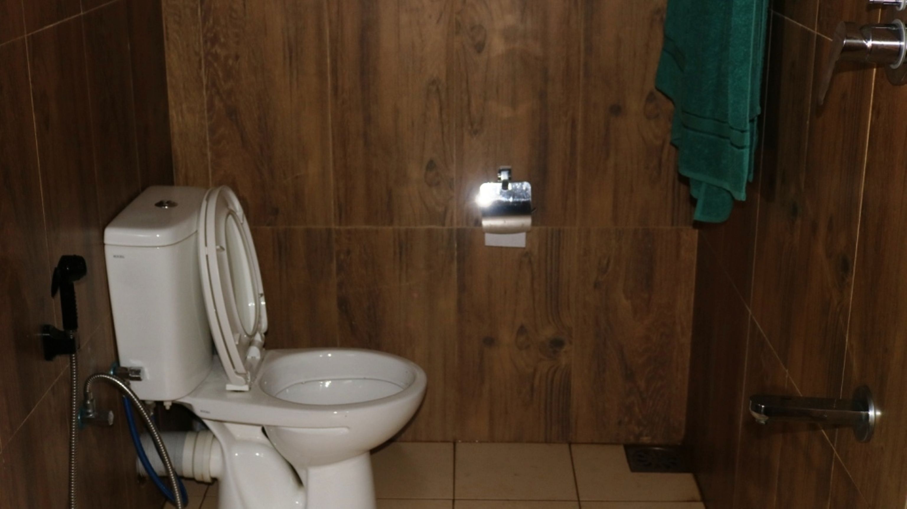 Un baño con inodoro blanco, paredes de madera, una toalla verde colgada en la puerta y un dispensador de papel higiénico visible.