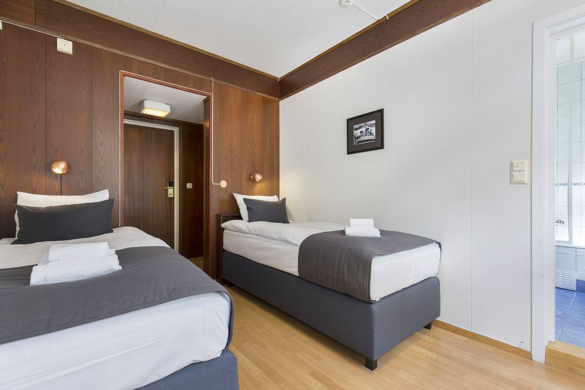 Une chambre d’hôtel avec deux lits, des lambris en bois, un tableau encadré au mur et une porte donnant sur une autre pièce.