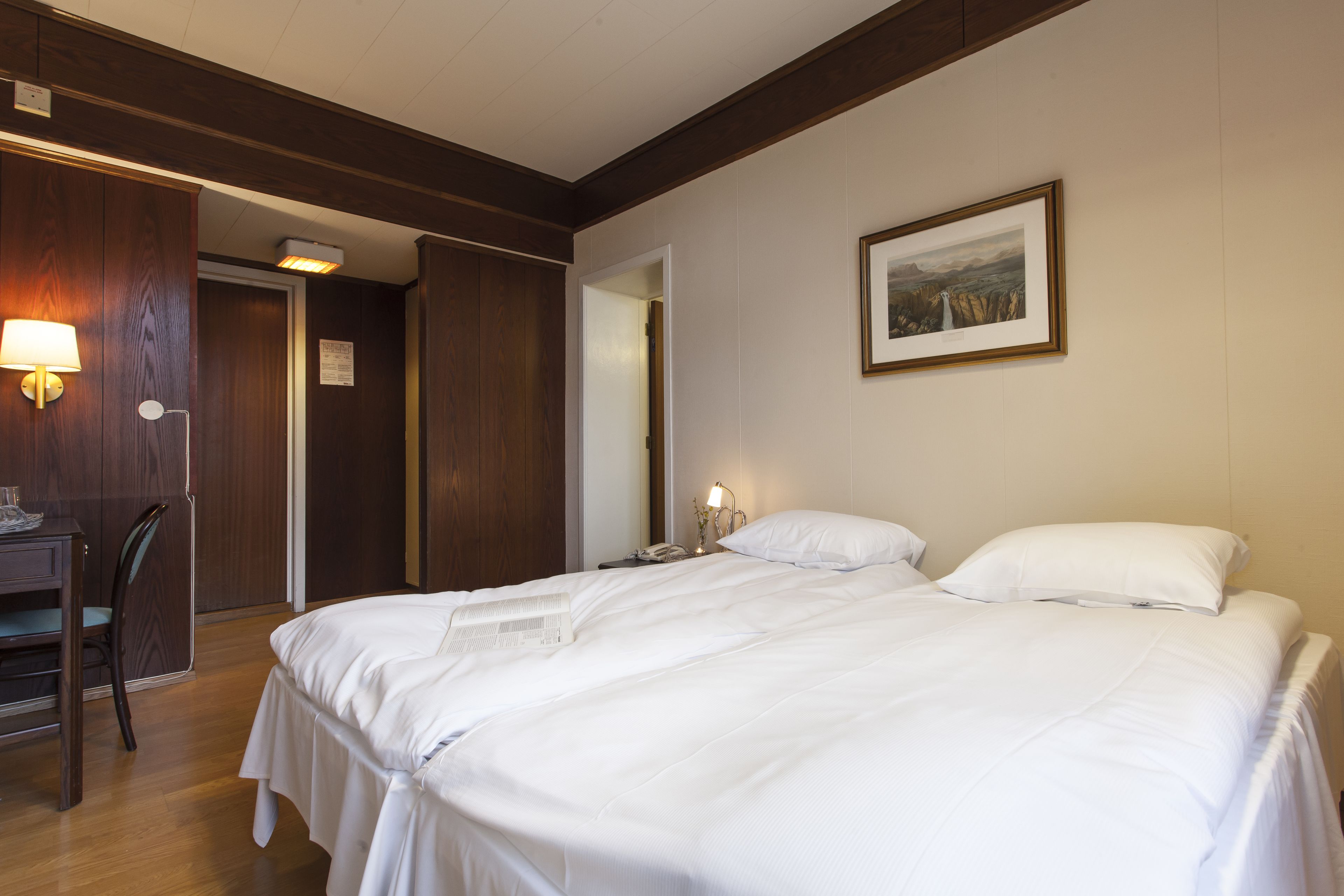 A hotel room with a large bed, a desk with a chair, a bedside table with a lamp, and a framed picture on the wall.