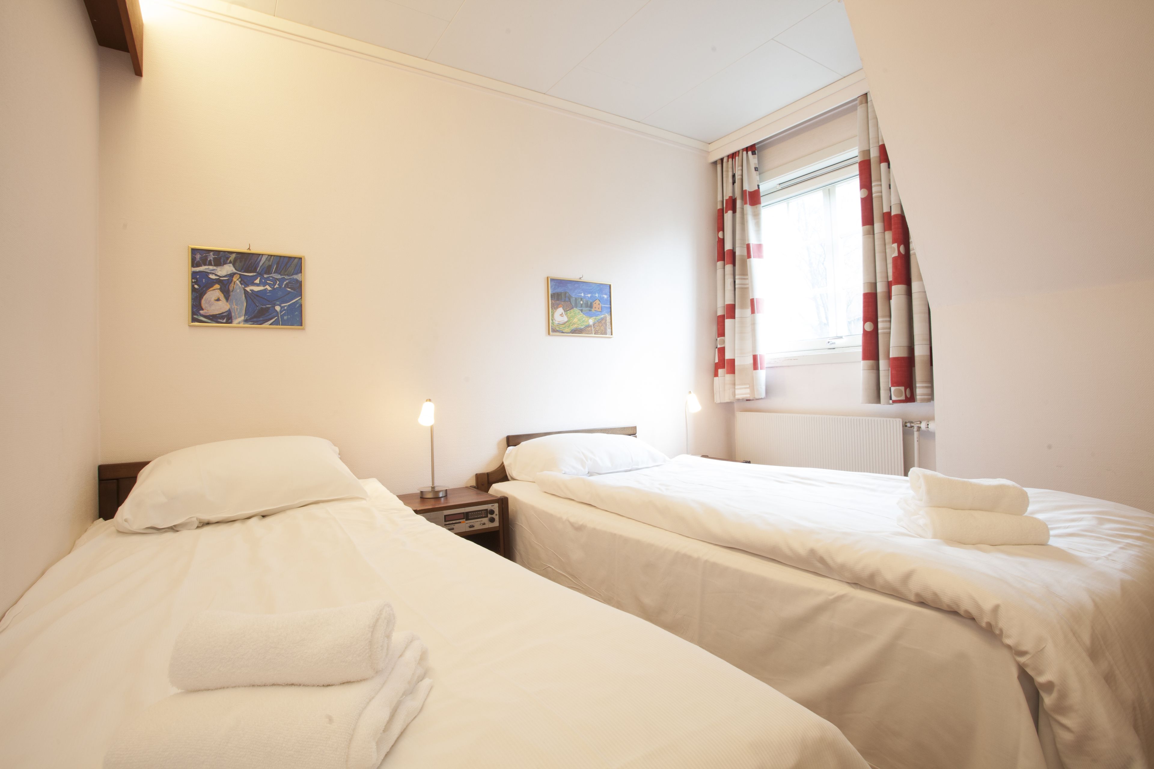 A hotel room with two beds, a nightstand with a lamp, and artwork on the walls.
