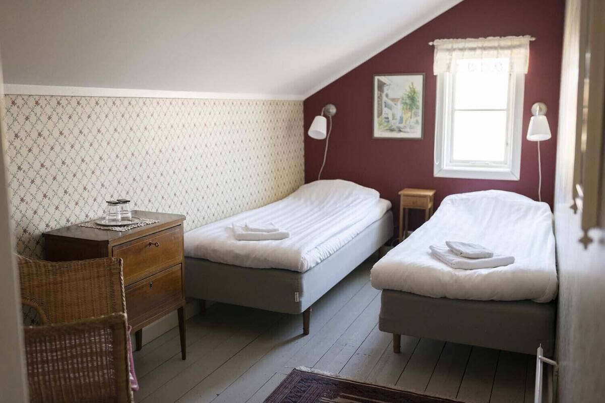 A bedroom with two single beds, a wooden dresser, a wicker chair, and a small table with a lamp.
