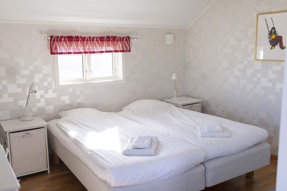 A bedroom with a bed, two bedside tables, a window with a red valance, and a wall-mounted air conditioner.