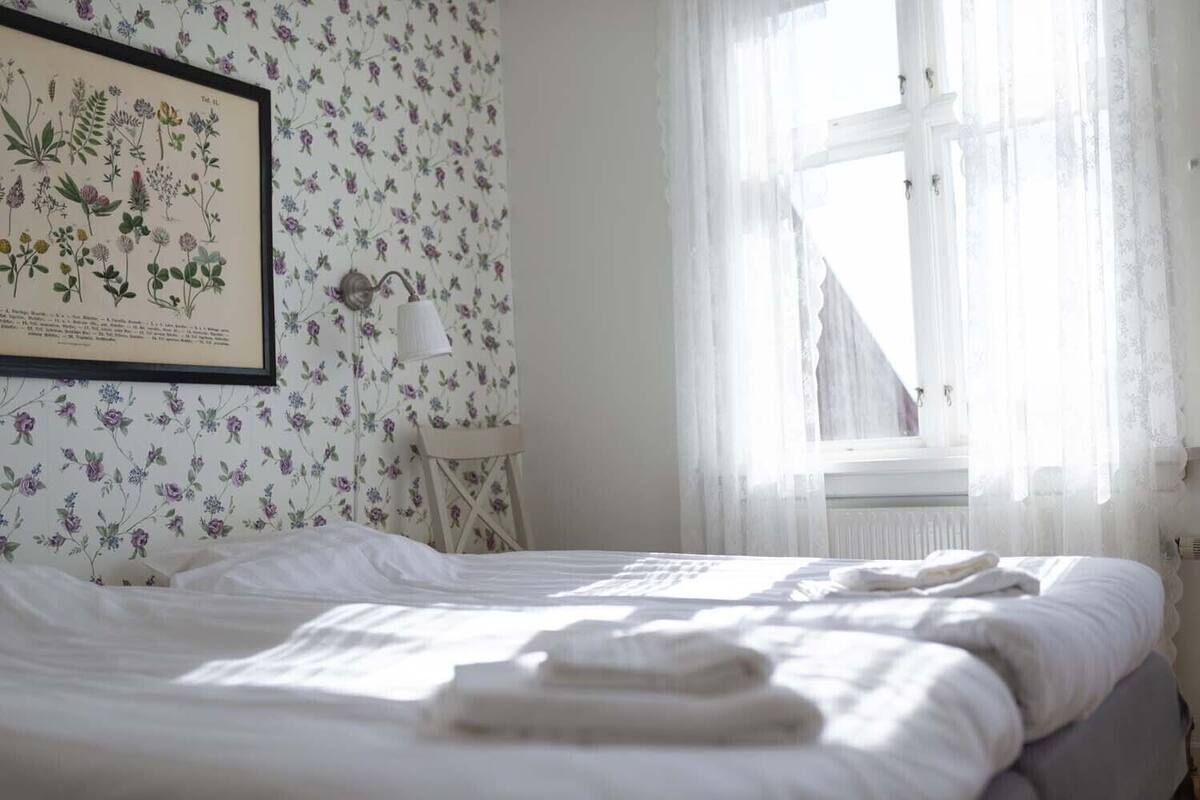 A bedroom with a floral wallpaper, a framed botanical illustration, a bedside lamp, and a window with sheer curtains.