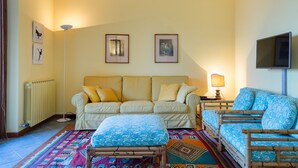 A living room with a beige sofa, a blue patterned sofa, a patterned ottoman, and a floor lamp. There are framed pictures on the wall and a television on the right. - Impero House Rent - Il Bosco (Stresa)