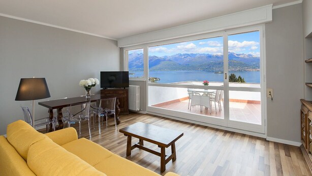 A living room with a yellow sofa, a wooden coffee table, a TV, and a dining table with chairs. There is a large window with a view of mountains and a lake. - Impero House Rent - Miralago (Stresa)
