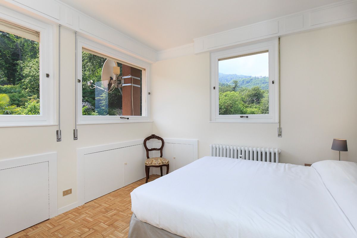 A bedroom with a bed, two windows, a chair, a radiator, and a lamp.