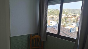 A corner room with a wooden chair, a small table, and a window overlooking a residential area with buildings and greenery. - Hotel Costa Andaluza (Motril)