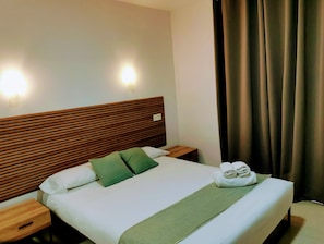 A neatly made bed with a wooden headboard, two pillows, a green throw blanket, and a set of towels on the side.