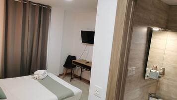 Habitación de hotel con cama, escritorio, silla, televisor y baño con lavabo y espejo.