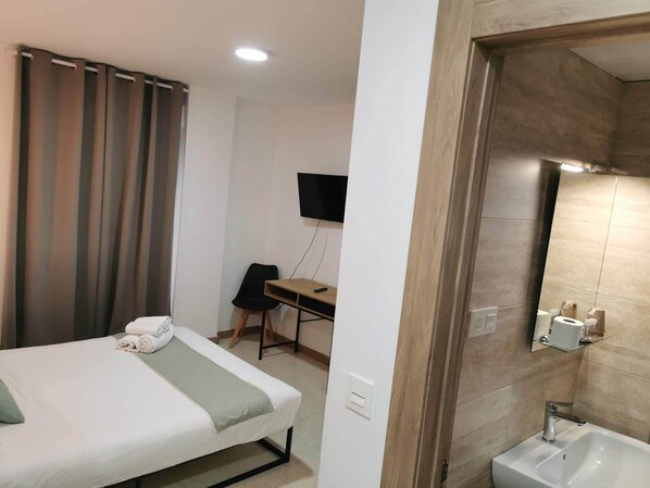 A hotel room with a bed, a desk, a chair, a TV, and a bathroom with a sink and mirror.