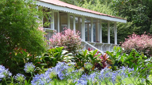 A white building with a porch surrounded by lush greenery and flowering plants. - Rondo Retreat Centre (Kakamega)