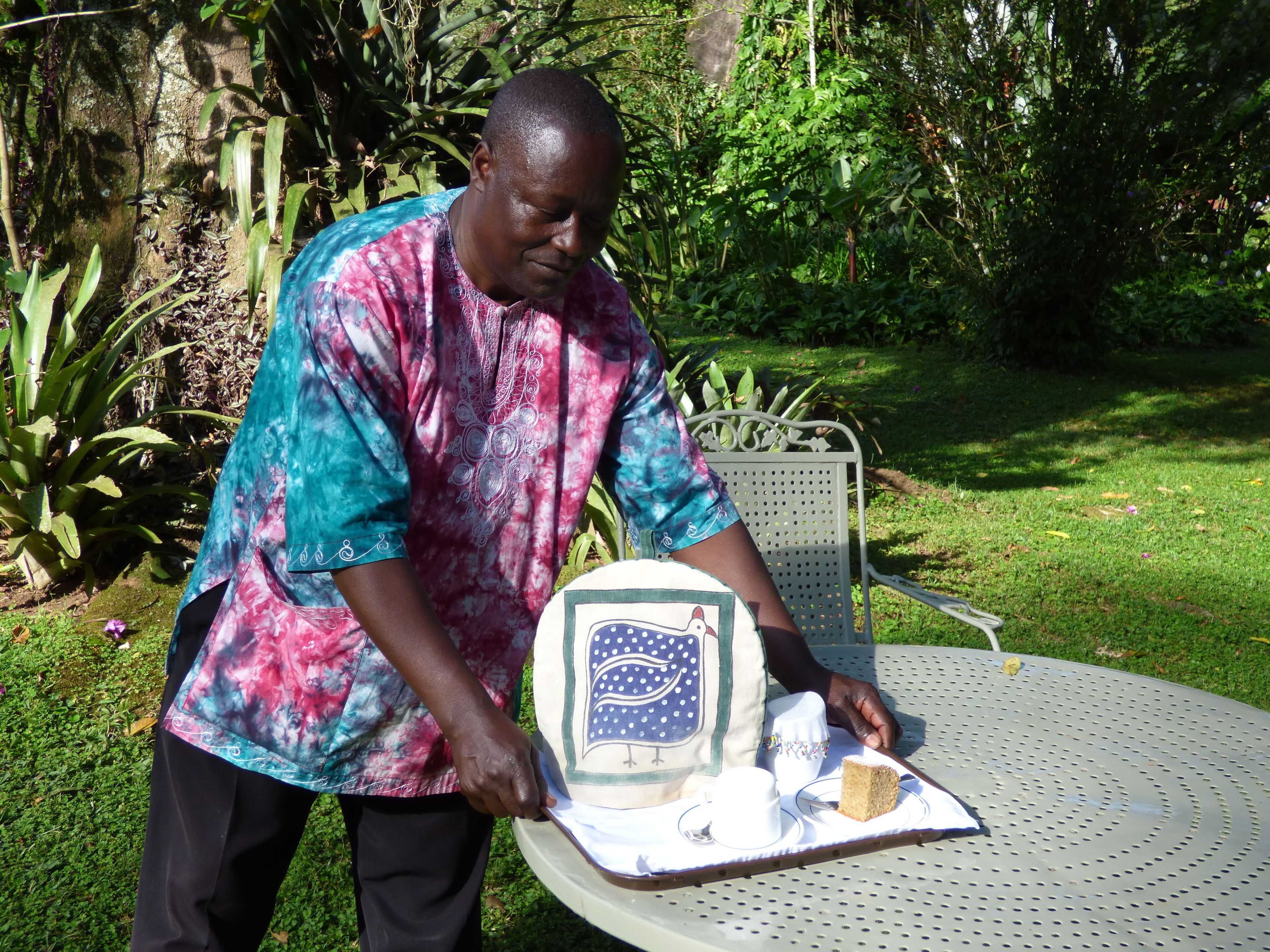 A person cutting a cake on a table outdoors with greenery in the background.