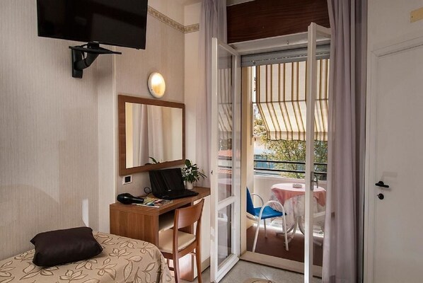 Habitación de hotel con cama, escritorio con laptop, silla, televisor montado en la pared y puerta corrediza de vidrio que da a un balcón exterior con mesa y sillas.