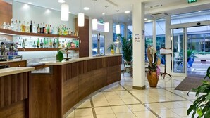 Un vestíbulo de hotel con una recepción, diversas botellas de licor en estantes y grandes ventanales que dejan entrar luz natural.