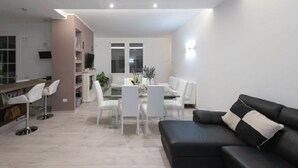 Ein modernes Wohnzimmer mit einer schwarzen Ledersofa, weißen Sesseln, einem Glastisch und einer Küchenzeile mit weißen Schränken und einer Bar.