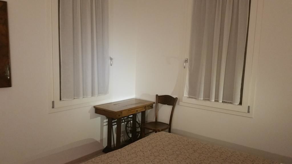 A small room with a single bed, a wooden desk, and a chair. There are two windows with white curtains.