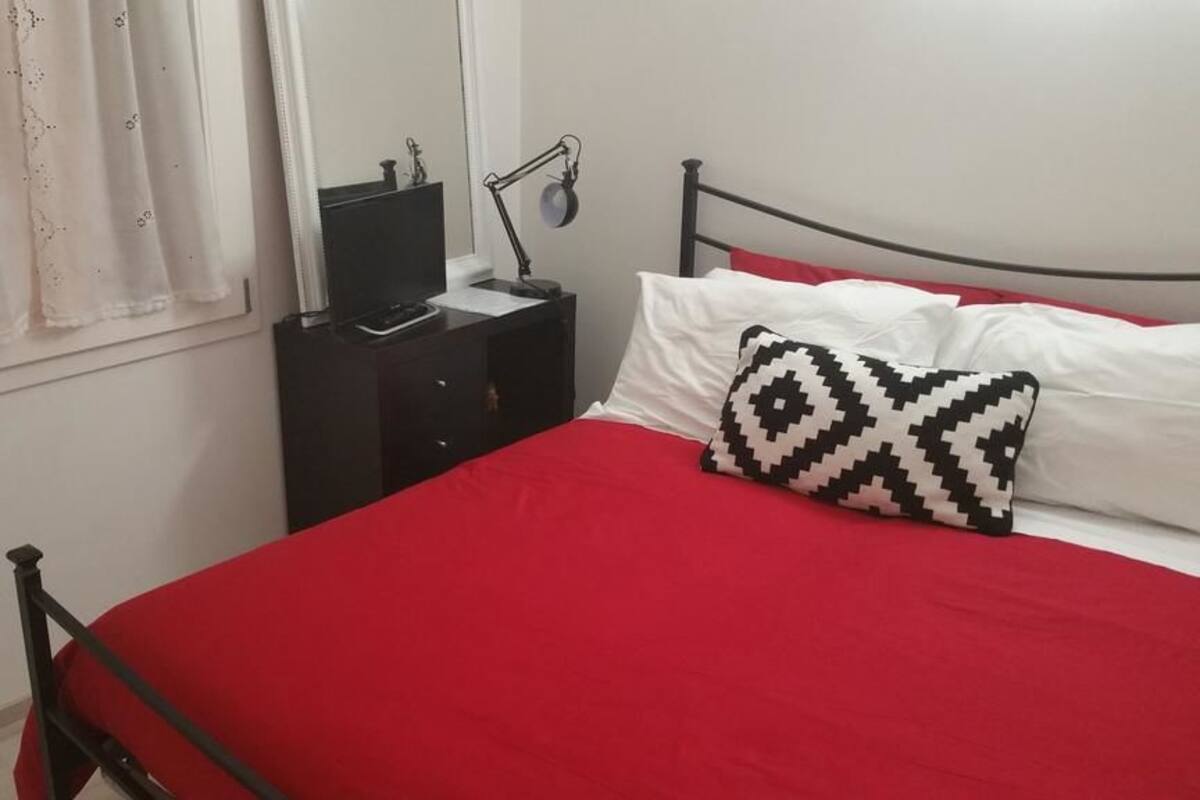 Una cama con colcha roja y almohada estampada en blanco y negro, una mesita de noche negra con una lámpara y una ventana con cortinas de gasa.