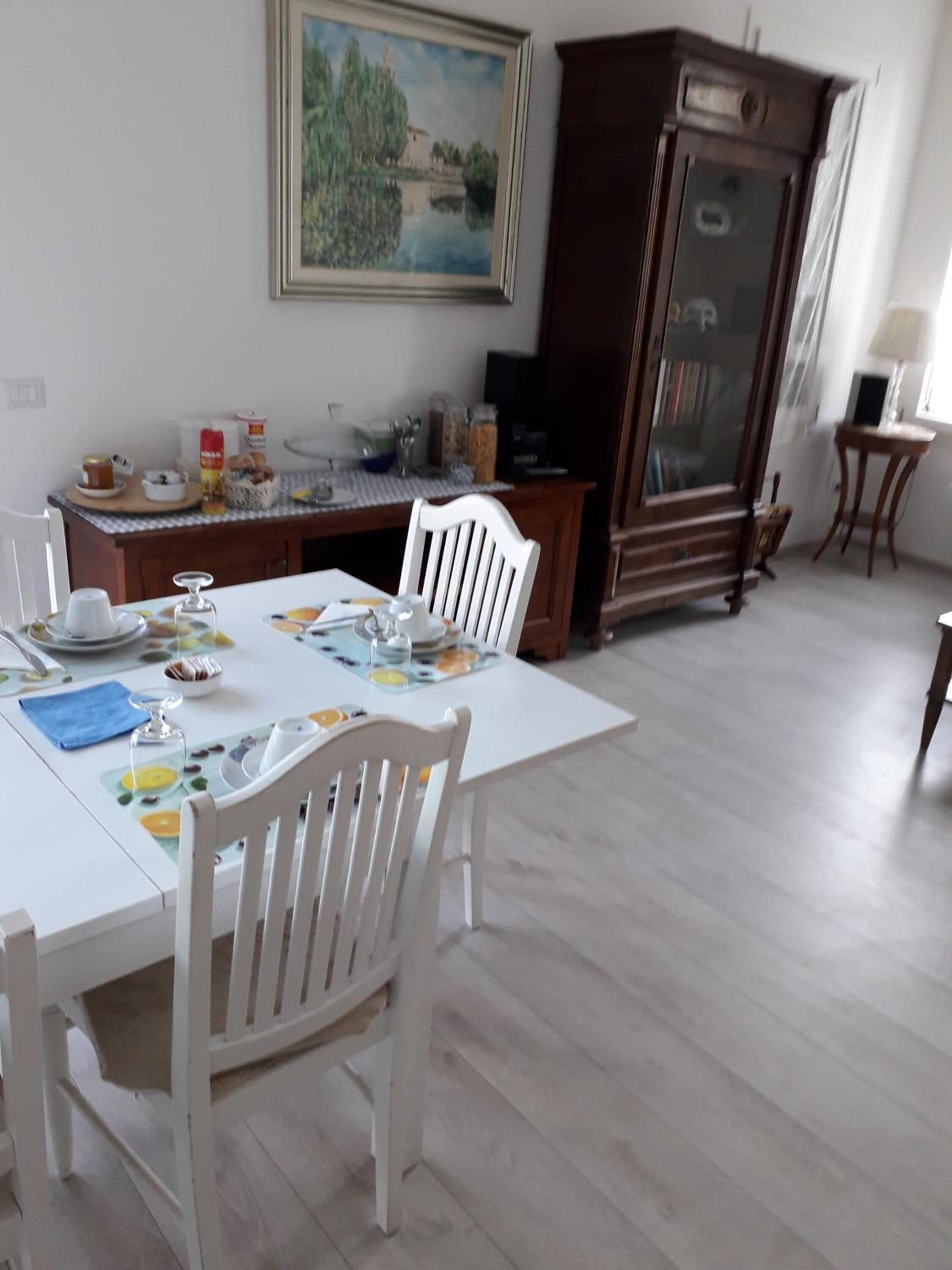 Un'area pranzo con un tavolo apparecchiato per quattro persone, sedie bianche, un mobiletto in legno e un quadro appeso al muro.