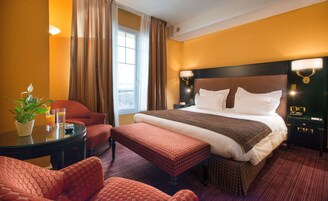 Quarto de hotel com uma cama grande, área de estar com mesa e cadeiras, planta em vaso e janela com cortinas.