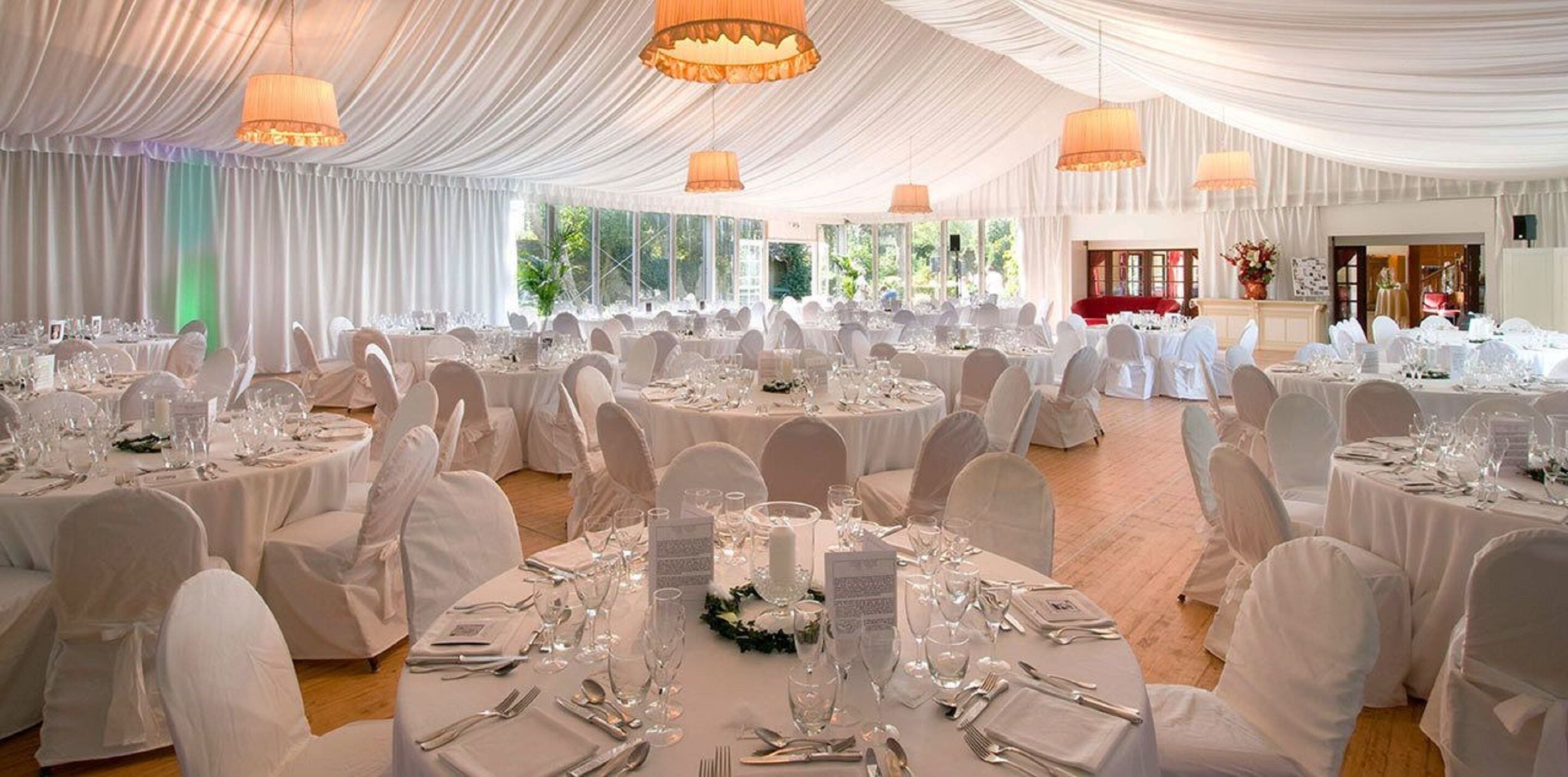 A banquet hall with round tables set for a formal event, featuring white tablecloths and chairs, and large windows providing natural light.