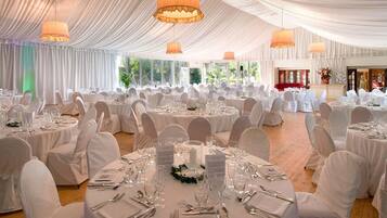 A banquet hall with round tables set for a formal event, featuring white tablecloths and chairs, and large windows providing natural light.