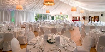 Un salón de banquetes con mesas redondas dispuestas para un evento formal, con manteles blancos y sillas, y ventanas amplias que dejan entrar luz natural.