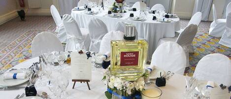 A banquet hall with round tables set for a formal event, featuring white tablecloths, glassware, and a bottle of 'HABIT ROUGE' perfume.