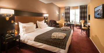 A hotel room with a large bed, a desk with a telephone, a chair, a small table with a plant, and a television on the wall.