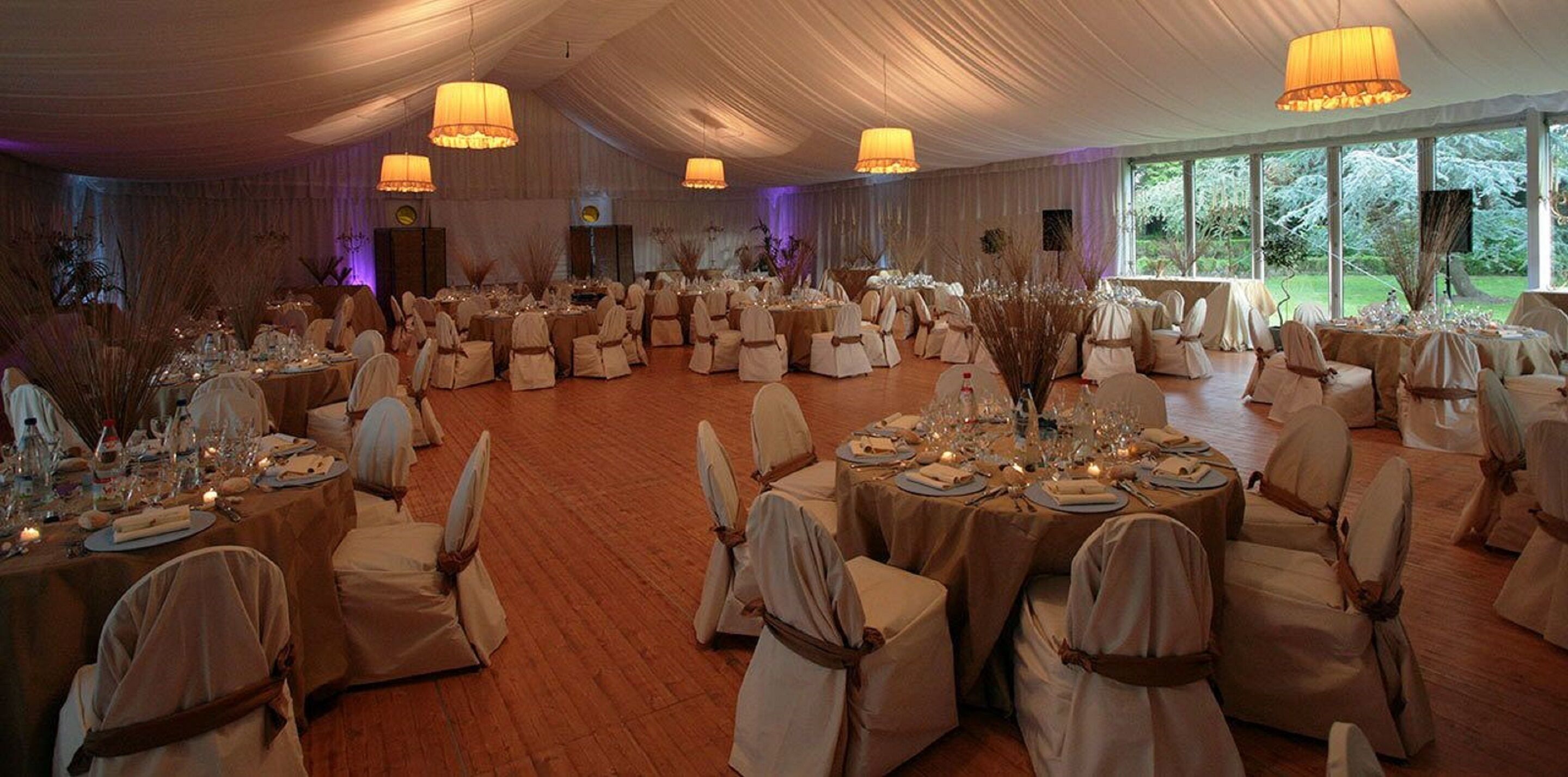 A banquet hall with round tables set for a formal event, featuring white tablecloths, chairs with brown sashes, and ambient lighting.