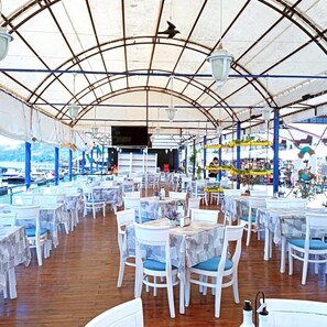 An outdoor restaurant with white tables and chairs, a covered roof, and a view of the sea. - Lotos Hotel (Balchik)