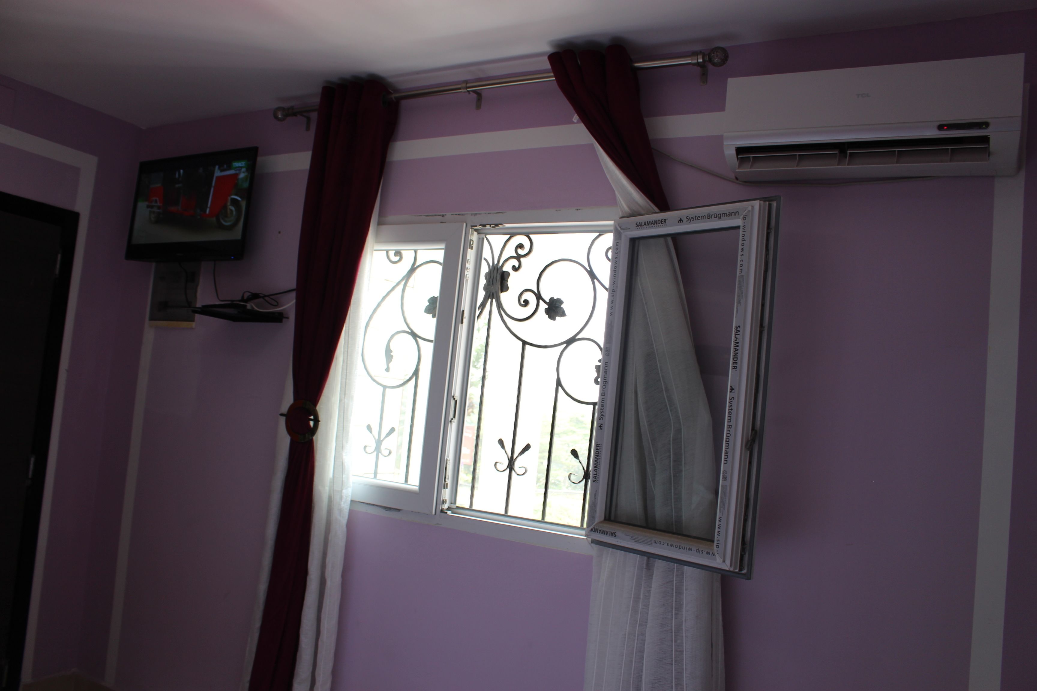 Une pièce aux murs violets, avec un téléviseur fixé au mur, un appareil de climatisation et une fenêtre ornée de barreaux en fer décoratifs.