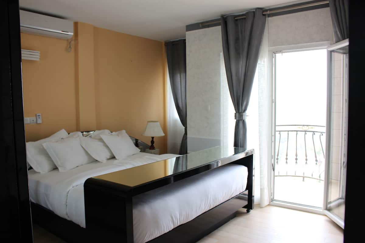 Quarto de hotel com uma cama grande, uma mesinha de cabeceira, uma luminária e vista para a varanda externa.
