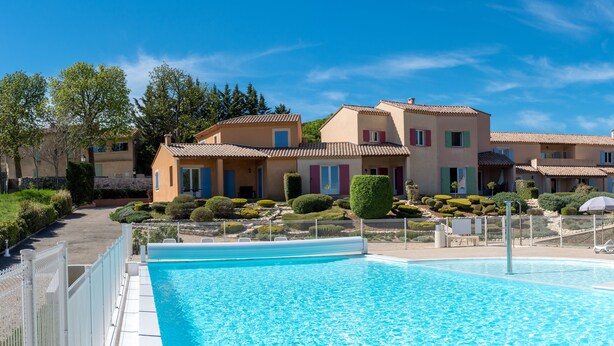 Une zone de piscine avec une piscine à l’eau d’un bleu limpide, entourée d’une clôture blanche. Au fond, on aperçoit des bâtiments avec des volets rouges et bleus, et un aménagement paysager bien entretenu.