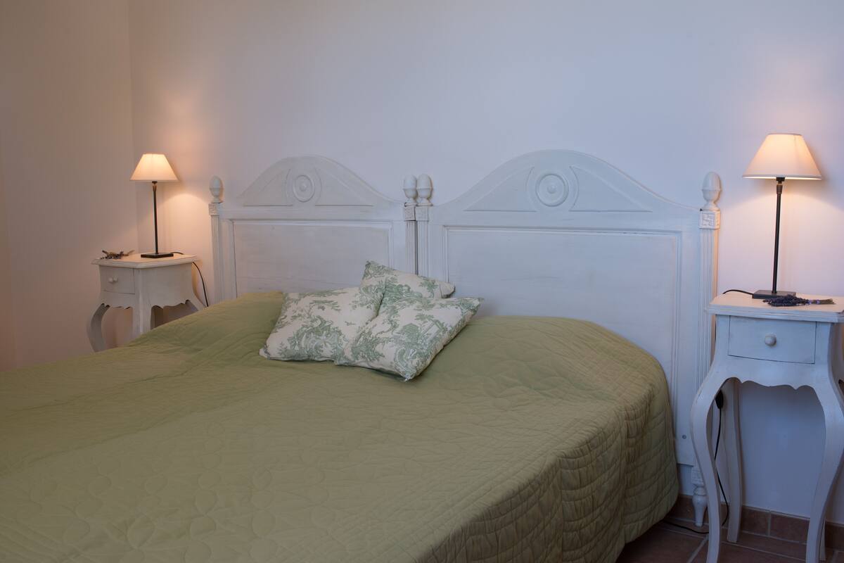 A neatly made bed with a green bedspread and two decorative pillows, flanked by two bedside tables with lamps, in a simple room with white walls and a classic headboard.