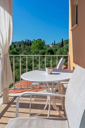 A balcony with a white table and chairs, a potted plant, and a view of greenery and hills. - Kapella Apartments (Corfu)