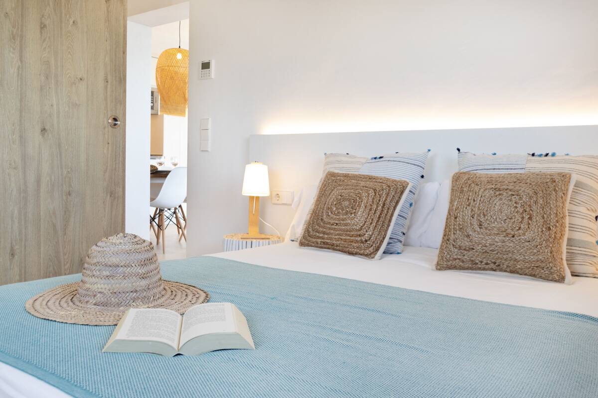 Una cama bien hecha con una colcha azul, un sombrero de paja y un libro abierto sobre la cama.